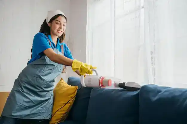 Smiling housekeeper vacuuming sofa for a clean and dust-free living room environment."