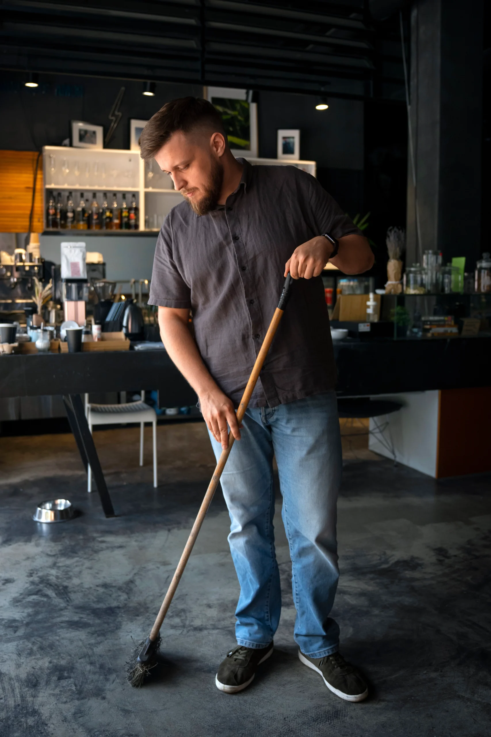 Man in gray shirt and blue jeans sweeping the floor of a dark cafe.