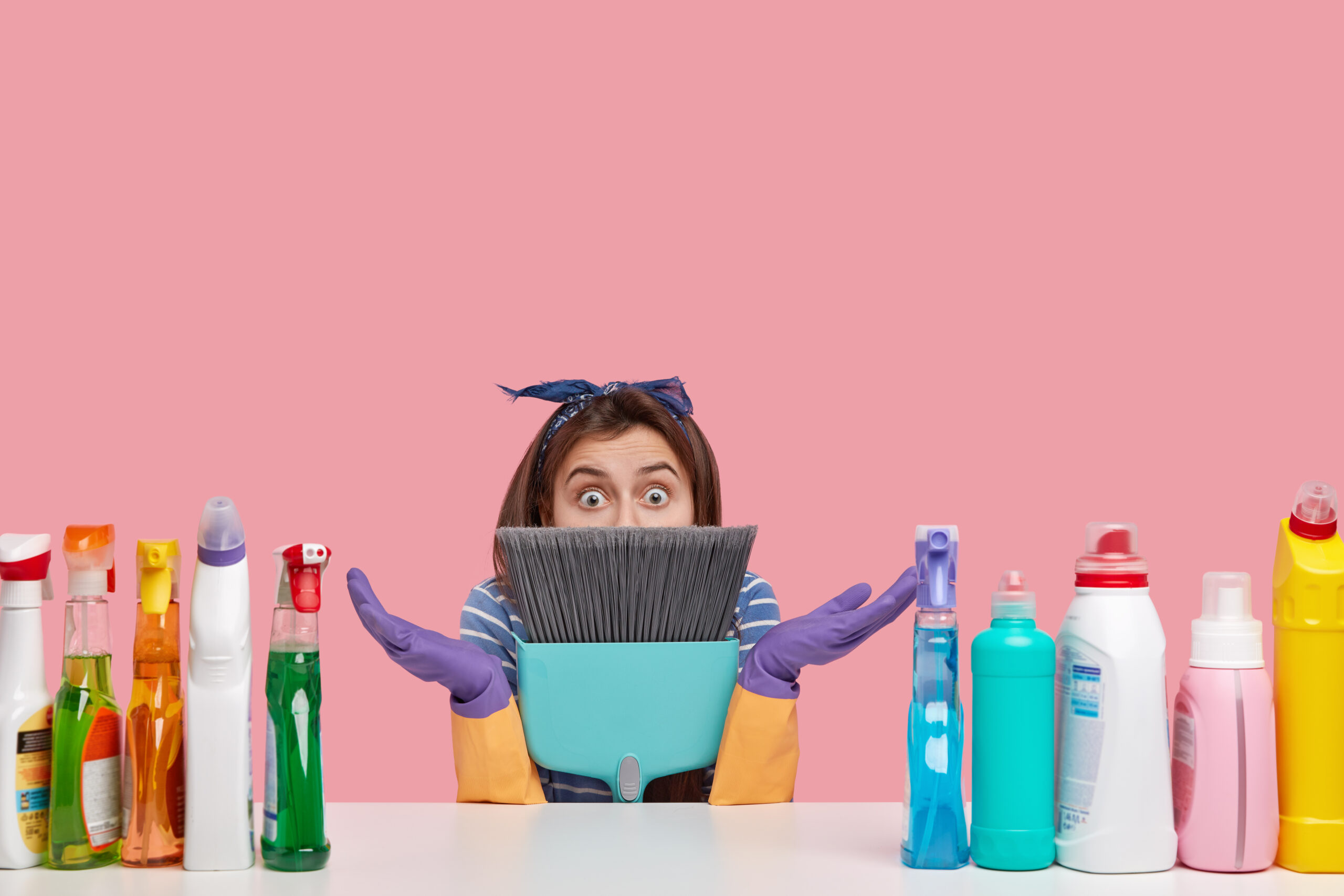 Cleaning Cost Calculation, Woman wearing cleaning gloves and a headband, holding a dustpan and broom, looking surprised in front of various cleaning supplies.