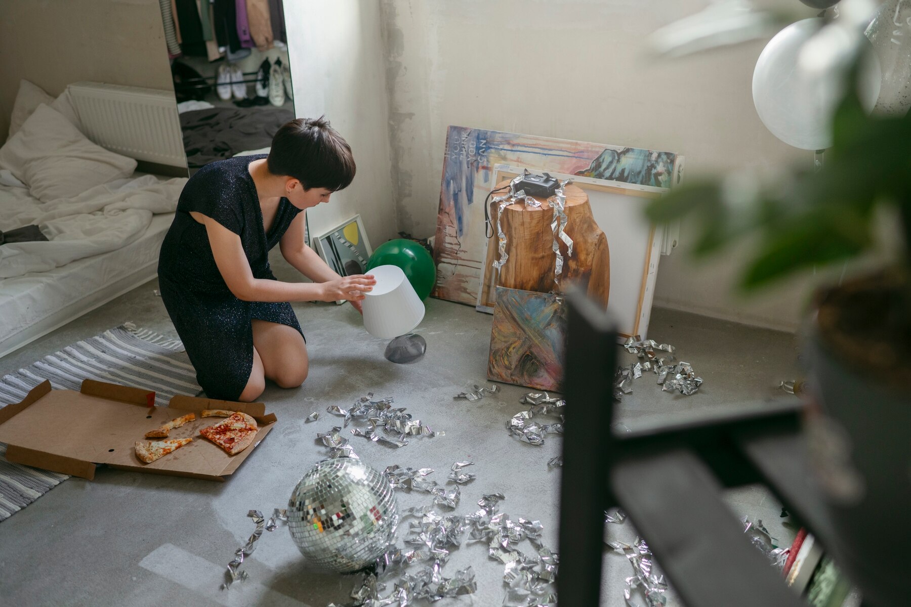 Woman kneeling on floor amidst party debris, arranging a white decorative bowl amongst art supplies and pizza boxes.