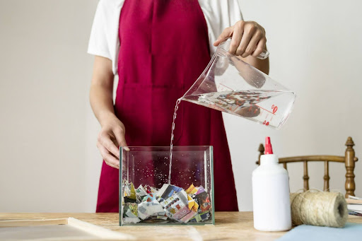 Person pouring water into a glass box filled with colorful paper scraps.