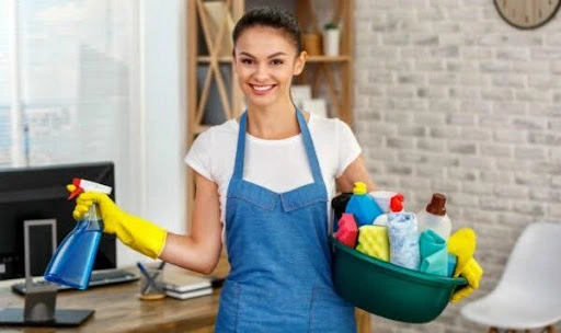 General cleaning and deep cleaning differences, miling woman in a blue apron holding cleaning supplies, including a spray bottle and a cleaning basket.