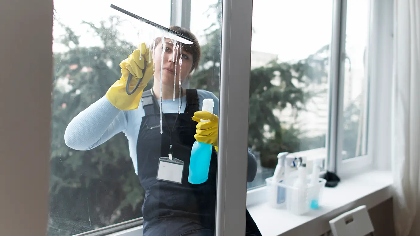 professional-window-cleaning-service CRmaids professional window cleaner wearing yellow gloves and overalls uses a squeegee and blue cleaning solution to clean a large indoor window, with cleaning supplies placed on the windowsill and trees visible outside.